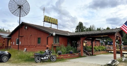 A rustic wooden building with a sign reading "Turtle Lodge," a motorcycle parked outside, and an American flag nearby.