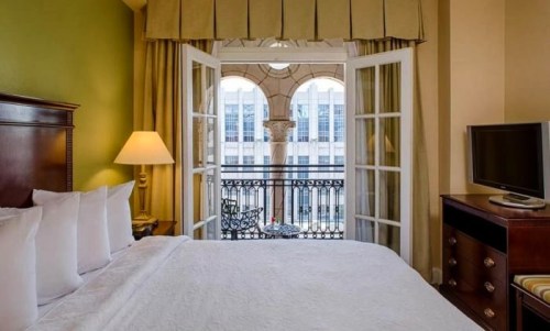 Cozy hotel room with a bed, lamp, and TV, featuring large windows opening to a balcony with a city view.