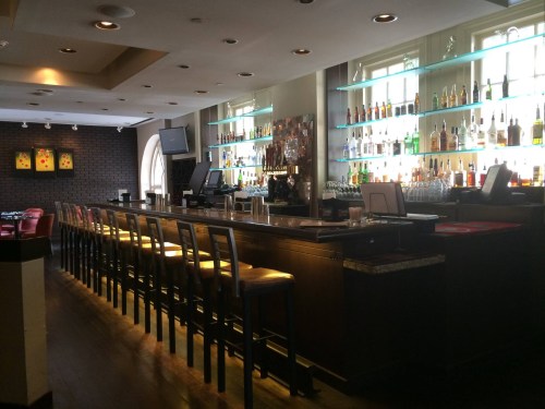 A modern bar with a long counter, illuminated shelves of liquor, and a cozy seating area in the background.