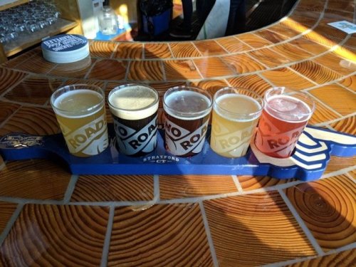 Five colorful beer samples arranged on a blue paddle, set against a wooden bar countertop.