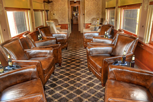 Interior of a vintage train car featuring leather armchairs, a patterned carpet, and small tables with drinks.