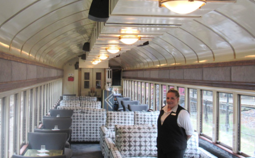 A smiling staff member stands in a vintage train car, featuring plush seating and large windows.