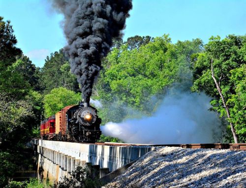A vintage steam train billows black smoke as it crosses a bridge surrounded by lush greenery.