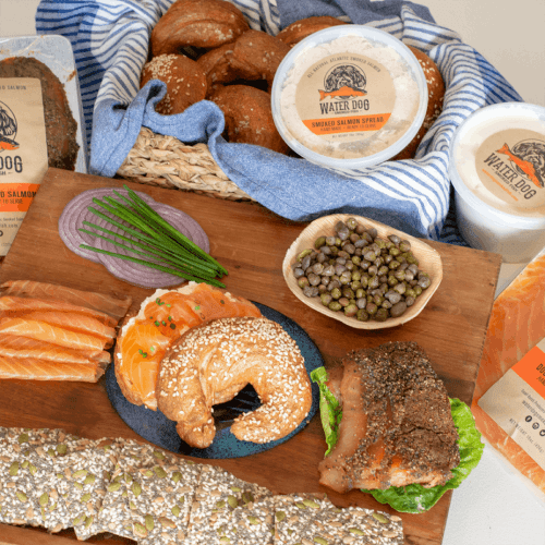 A spread of smoked salmon, bagels, capers, and a salmon spread, arranged on a wooden board with a blue cloth.
