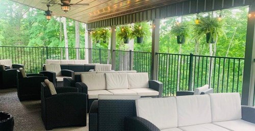 A cozy outdoor seating area with white sofas and green plants, surrounded by trees and a railing.