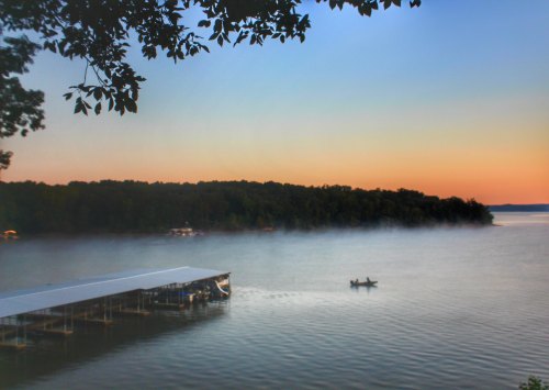 A serene lake at dawn with mist rising, a dock in the foreground, and a small boat on the water.