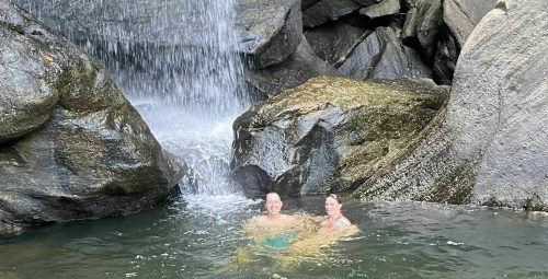 Two people smiling in a natural pool beneath a waterfall, surrounded by large rocks.