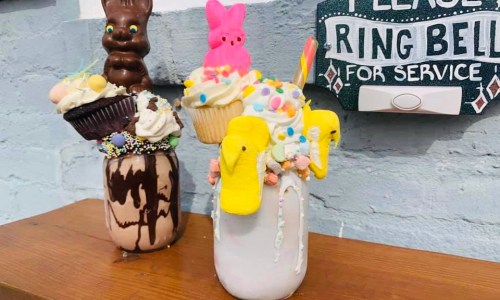 Two colorful milkshakes topped with cupcakes, candy, and marshmallow treats, set against a rustic background.