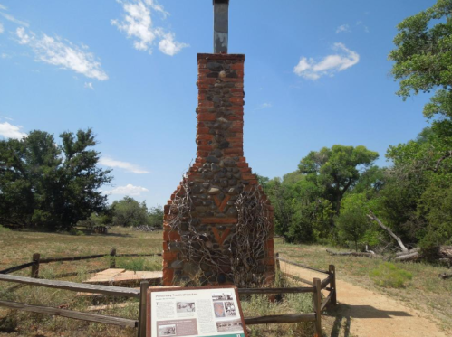 A stone and brick chimney structure with a sign nearby, set in a grassy area under a blue sky with scattered clouds.