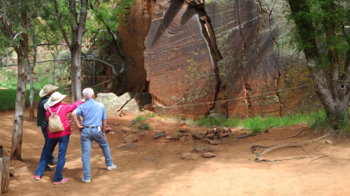 Two people observe rock formations in a natural setting, surrounded by trees and red soil.