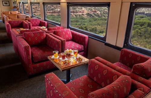 Cozy train lounge with red patterned seating and a table of drinks and snacks, overlooking a scenic landscape.