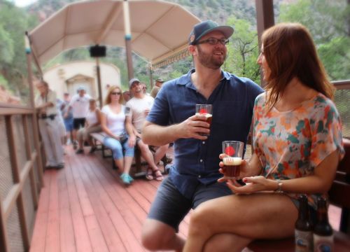 A couple enjoys drinks on a train, surrounded by other passengers in a scenic outdoor setting.