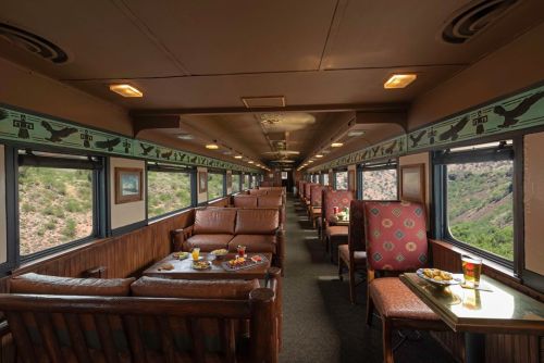 Interior of a train car featuring comfortable seating, tables with snacks, and large windows showcasing a scenic view.