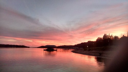 A serene lake at sunset, with vibrant pink and orange skies reflecting on the water and a dock in the foreground.