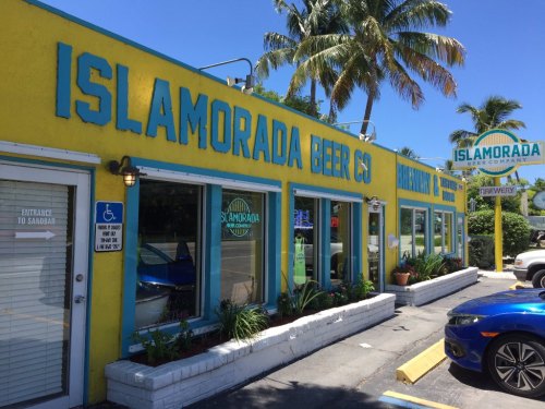 Exterior of Islamorada Beer Company, featuring bright yellow walls, blue trim, and palm trees in a sunny setting.
