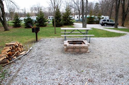A campsite with a picnic table, fire pit, stacked firewood, and gravel ground, surrounded by trees and RVs in the background.
