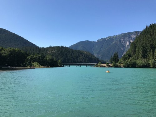 A serene lake surrounded by mountains and lush trees, with a bridge in the distance under a clear blue sky.