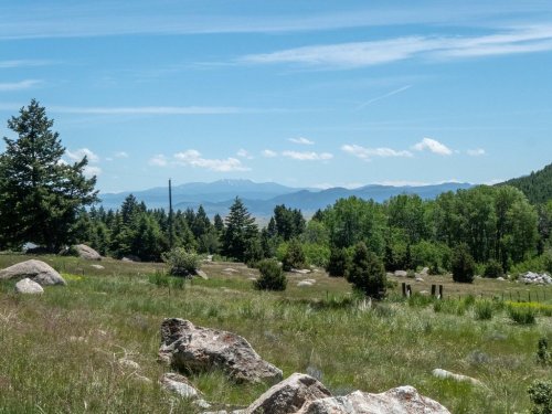 A scenic view of rolling hills, trees, and distant mountains under a clear blue sky with scattered clouds.