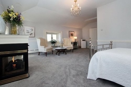 Cozy bedroom with a fireplace, two armchairs, a small table, and soft gray carpet, featuring elegant decor and natural light.