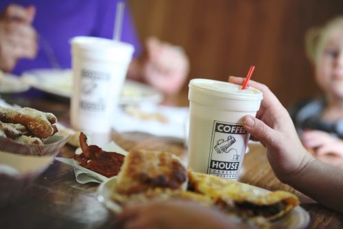 A hand holds a coffee cup at a table with food, including pastries and a plate of breakfast items.