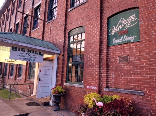 Exterior of a brick building with a sign for "Big Mill Co." and "Vintage 1889 Casual Dining," featuring colorful plants.