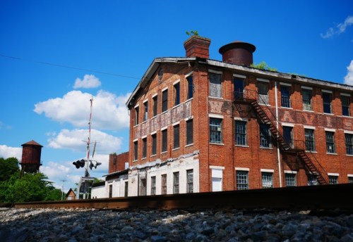 An old brick building with a fire escape, water tower, and blue sky, viewed from a railway track.