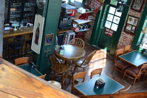 Cozy restaurant interior with wooden tables, chairs, and a bar area, featuring green walls and warm lighting.