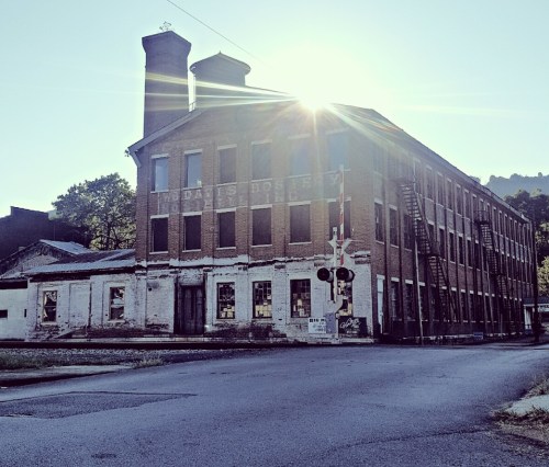 Abandoned brick building with two smokestacks, sunlight shining behind it, near a railroad crossing.