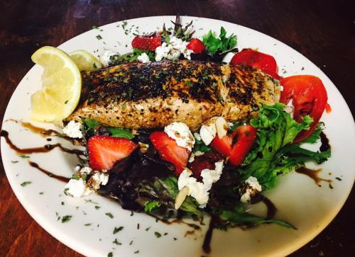 Grilled salmon on a bed of mixed greens, topped with strawberries, tomatoes, goat cheese, and lemon slices.