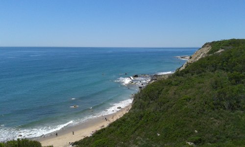A scenic view of a beach with gentle waves, rocky shoreline, and lush green hills under a clear blue sky.