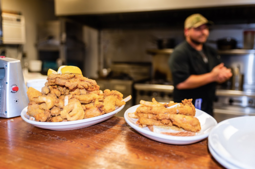 A wooden table with two plates of fried chicken, one with a lemon slice, and a chef in the background.