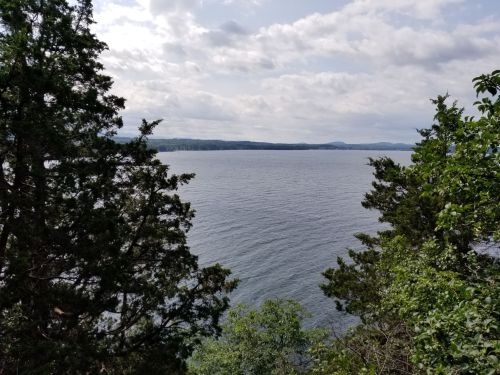 A serene view of a lake surrounded by trees under a cloudy sky, with distant hills on the horizon.