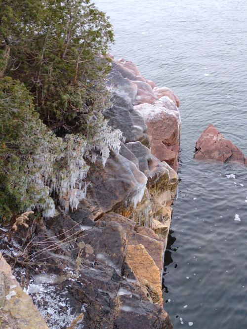 Rocky shoreline with icicles hanging from the edge, surrounded by calm water and greenery.