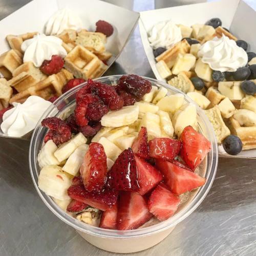 A cup of yogurt topped with sliced bananas, strawberries, and raspberries, with two plates of waffles in the background.