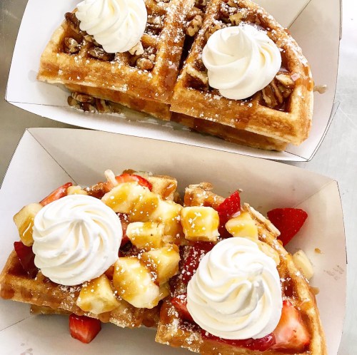Two delicious waffles topped with whipped cream, strawberries, bananas, and drizzled with syrup, served in paper trays.