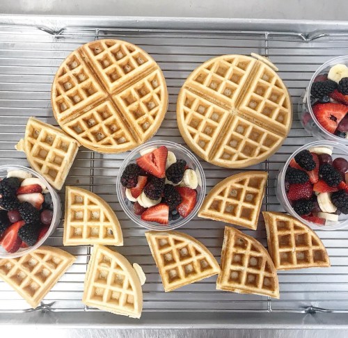 Two golden waffles on a cooling rack, surrounded by cups of mixed berries and sliced bananas.