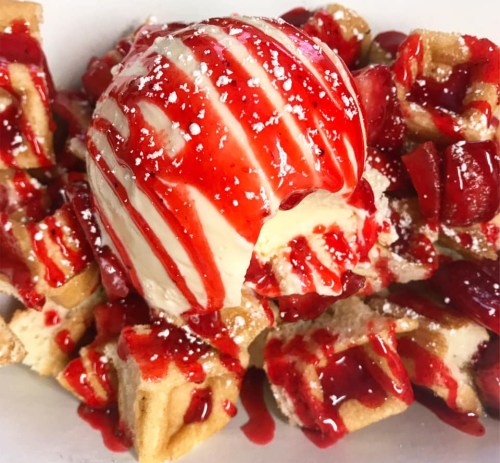 A bowl of waffle pieces topped with vanilla ice cream, strawberry sauce, and powdered sugar.