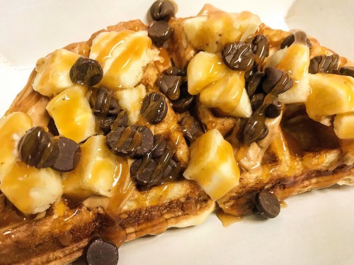 A close-up of a waffle topped with banana slices, chocolate chips, and a drizzle of peanut butter.