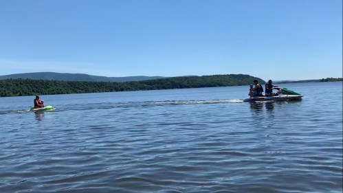A jet ski with two people rides across a calm lake, surrounded by green hills under a clear blue sky.