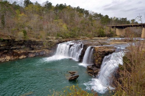 A serene waterfall cascades into a turquoise pool, surrounded by lush greenery and rocky cliffs under a cloudy sky.