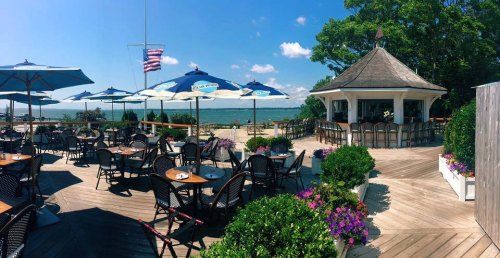 Outdoor dining area with tables, umbrellas, and a gazebo overlooking a scenic waterfront. Bright blue sky and greenery.
