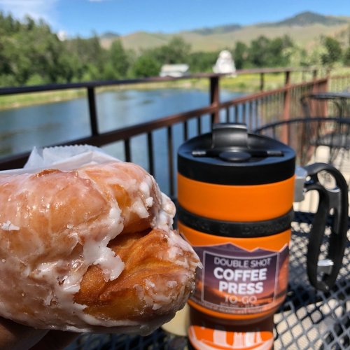 A glazed donut in hand with a coffee press and scenic river view in the background.