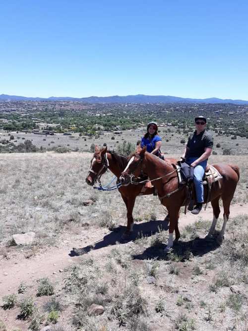 Two people on horseback in a scenic landscape with mountains in the background and clear blue skies.