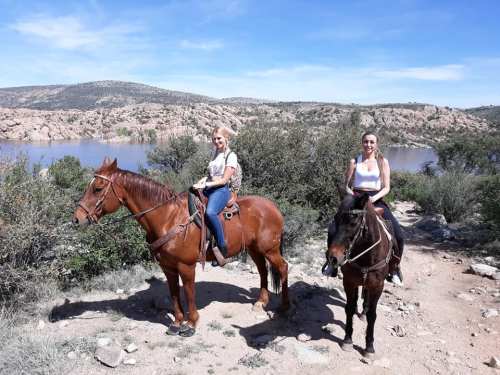 Two women riding horses along a scenic trail by a lake, surrounded by hills and greenery under a clear blue sky.