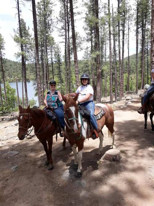 Two women on horseback in a forested area, with trees and a lake visible in the background.