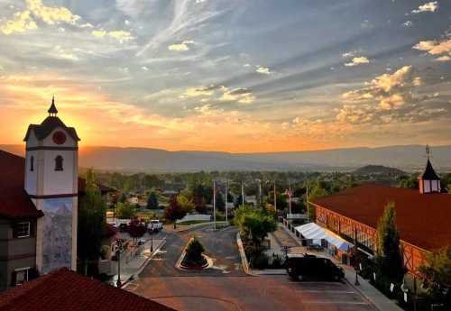 A scenic sunset view over a town, featuring buildings, trees, and mountains in the background under a colorful sky.