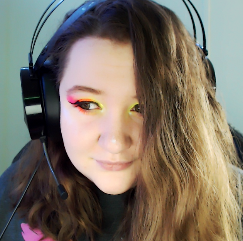 A person with long, wavy hair wearing a headset and colorful eye makeup, looking at the camera with a slight smile.