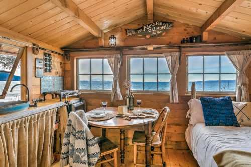 Cozy cabin interior with a dining table, bed, and large windows overlooking a serene lake view. Warm wooden decor.