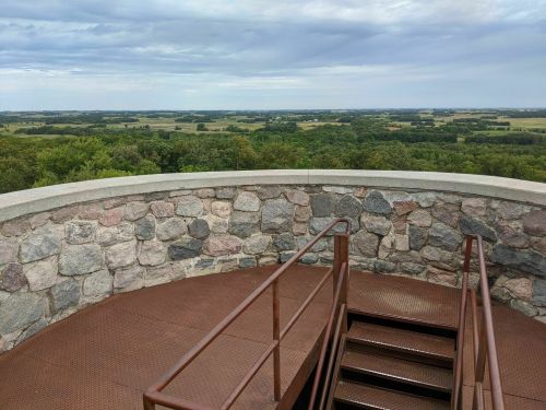 View from a stone observation deck overlooking lush green fields and a cloudy sky. Stairs lead down from the platform.