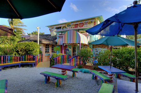 Colorful outdoor seating area with striped tables and umbrellas, surrounded by lush greenery and a vibrant building in the background.